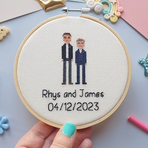 Puede incluir: Un bordado de punto de cruz en un aro de madera. El dise&ntilde;o presenta dos figuras con trajes, con los nombres "Rhys and James" y la fecha "04/12/2023" bordados debajo. El fondo es de tela beige claro.