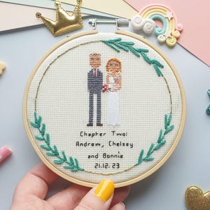 Puede incluir: Bordado de punto de cruz en un aro de madera. El dise&ntilde;o presenta una pareja con atuendo de boda, rodeada de hojas verdes y un borde dorado. El texto dice "Chapter Two: Andrew, Chelsey and Bonnie 21.12.23". Una corona dorada y un adorno de arco&iacute;ris en el fondo.