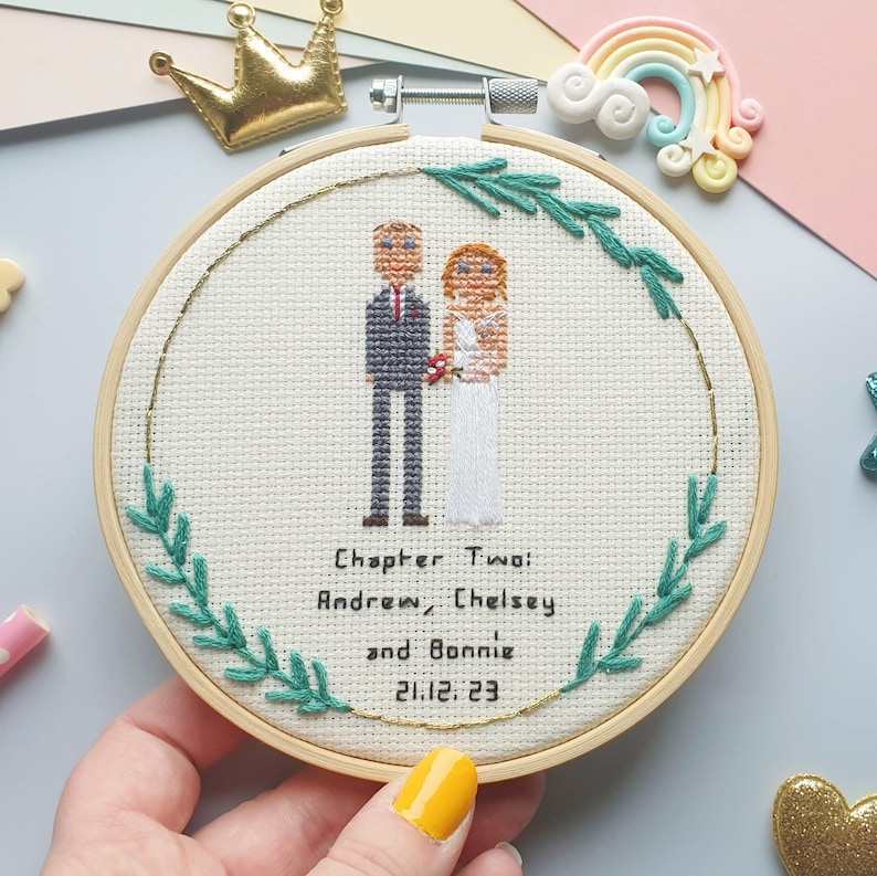 Puede incluir: Bordado de punto de cruz en un aro de madera. El dise&ntilde;o presenta una pareja con atuendo de boda, rodeada de hojas verdes y un borde dorado. El texto dice "Chapter Two: Andrew, Chelsey and Bonnie 21.12.23". Una corona dorada y un adorno de arco&iacute;ris en el fondo.