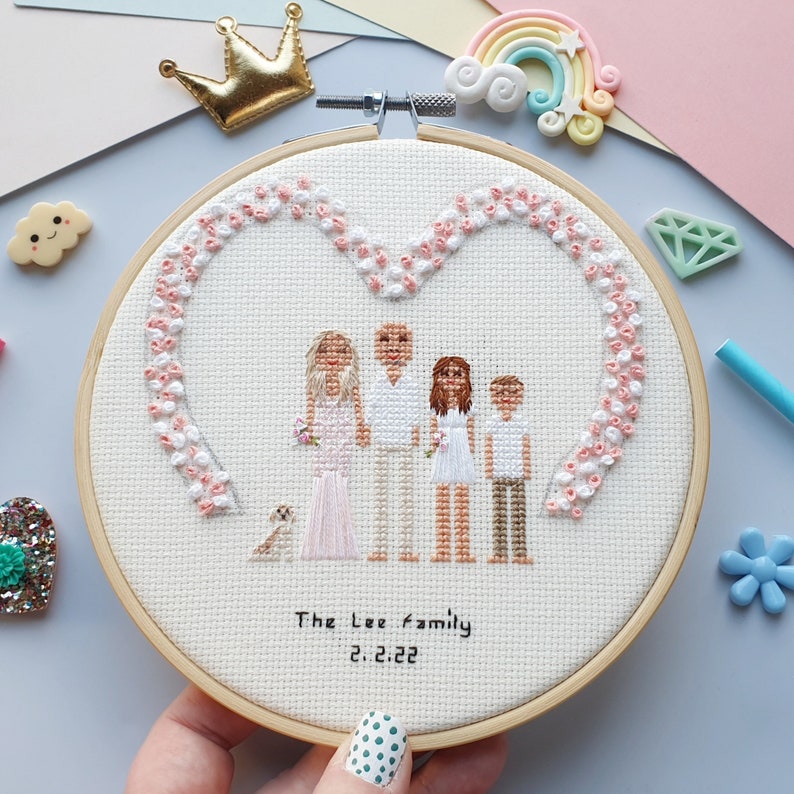 Puede incluir: Un retrato familiar bordado a punto de cruz en un bastidor de madera. El dise&ntilde;o presenta una familia de cuatro miembros y un perro peque&ntilde;o, enmarcado por un borde en forma de coraz&oacute;n de flores rosas y blancas. El texto "The Lee family 2.2.22" est&aacute; bordado debajo.