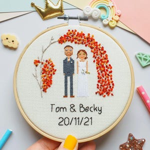 Puede incluir: Un bastidor de bordado de punto de cruz con una escena de boda con una novia y un novio bajo un arco de flores naranjas. Los nombres "Tom & Becky" y la fecha "20/11/21" est&aacute;n bordados debajo. El bastidor est&aacute; sobre un fondo colorido.