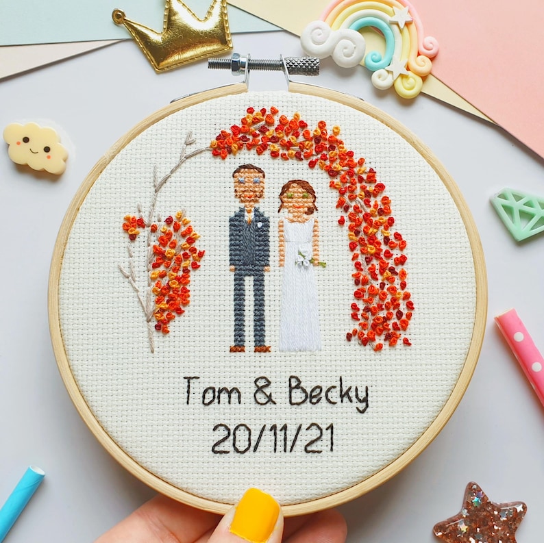 Puede incluir: Un bastidor de bordado de punto de cruz con una escena de boda con una novia y un novio bajo un arco de flores naranjas. Los nombres "Tom & Becky" y la fecha "20/11/21" est&aacute;n bordados debajo. El bastidor est&aacute; sobre un fondo colorido.