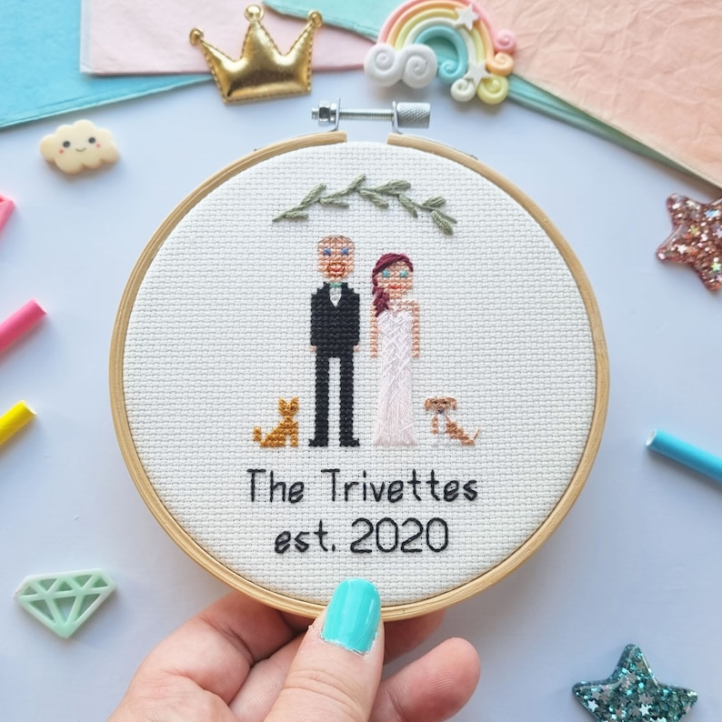 Puede incluir: Un bastidor de bordado de punto de cruz con una pareja, dos perros y el texto "The Trivettes est. 2020". La pareja viste ropa de boda. El bordado est&aacute; en un bastidor de madera, con un fondo de tela clara.