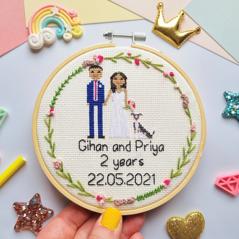 Puede incluir: Bordado de punto de cruz en un aro de madera con una pareja y un perro. El texto dice "Gihan and Priya 2 years 22.05.2021". El dise&ntilde;o est&aacute; rodeado por una corona floral. La pareja est&aacute; vestida con ropa de boda.