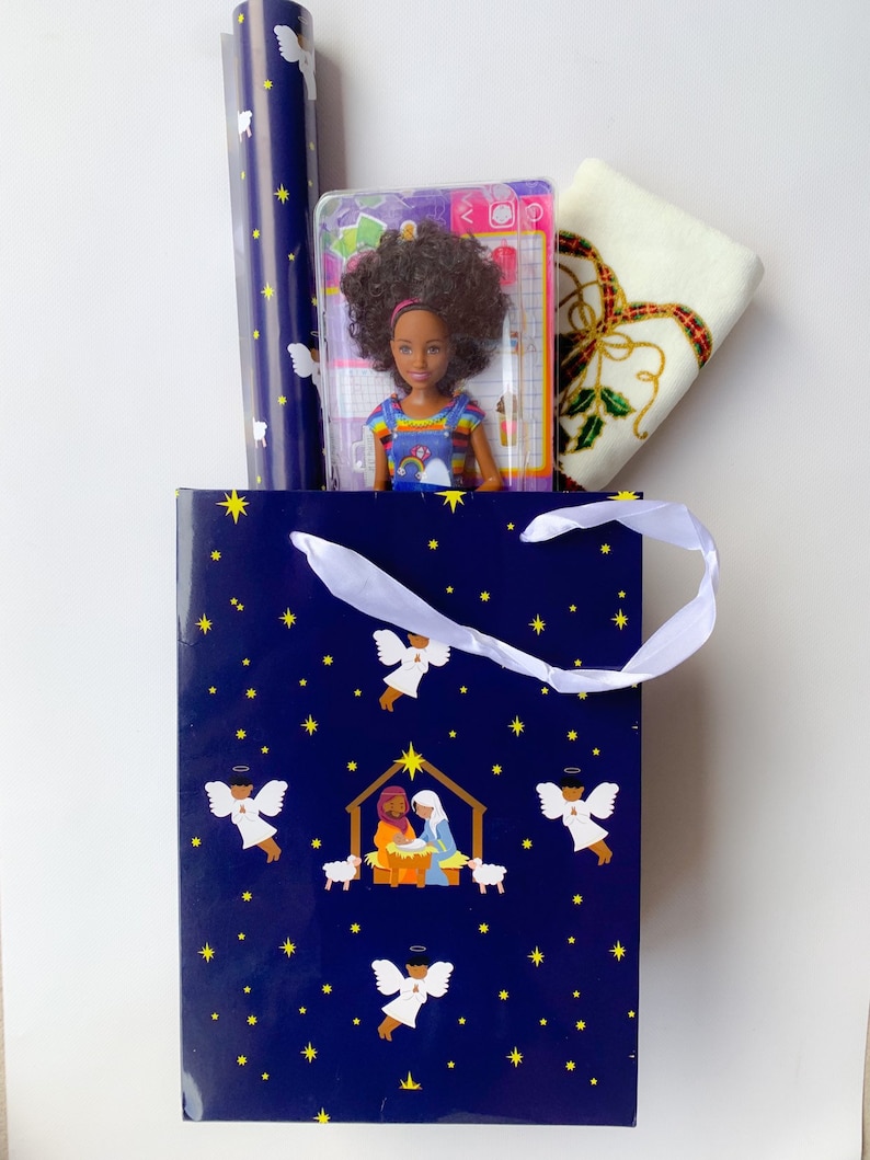 May include: A blue gift bag with a white ribbon and a nativity scene design. The bag contains a doll, wrapping paper, and a white towel with a gold and green design.