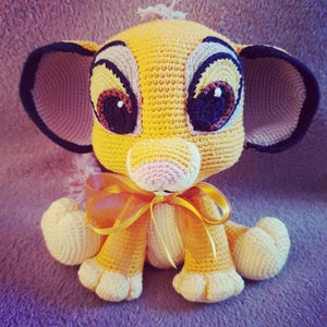 Könnte beinhalten: Ein gehäkeltes Stofftier-Löwenjunges mit großen Augen, einer rosa Nase und einer gelben und braunen Schleife. Das Löwenjunges sitzt auf einer violetten und grauen strukturierten Oberfläche.