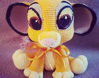 May include: A crocheted stuffed animal lion cub with large eyes, a pink nose, and a yellow and brown bow. The lion cub is sitting on a purple and gray textured surface.