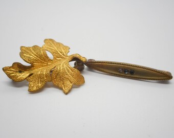 May include: A gold-toned leaf-shaped brooch with a long, thin, rectangular bar attached to the back. The bar has three small, clear rhinestones.