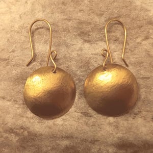 May include: A pair of gold-toned earrings with a round disc shape. The earrings have a hammered texture and a simple hook closure.