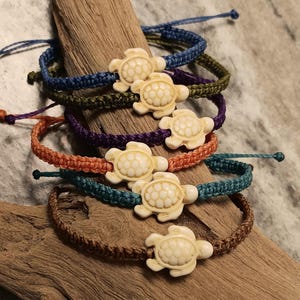 May include: A collection of five adjustable cord bracelets in blue, olive green, purple, orange, and brown. Each bracelet features a central cream-colored turtle charm with a detailed shell pattern, secured by a woven cord band with pull-string ends.