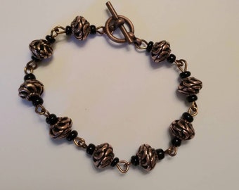 Könnte beinhalten: Ein Kupferarmband mit schwarzen Perlen und einem Toggle-Verschluss. Die Perlen haben ein strukturiertes, wirbelndes Design.
