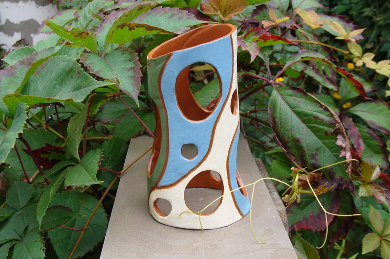K&ouml;nnte beinhalten: Eine handgefertigte Keramikvase mit einem einzigartigen, offenen Design. Die Vase hat eine Kombination aus Farben, darunter Hellblau, Creme und Braun, mit einer Reihe von kreisf&ouml;rmigen Aussparungen. Die Vase ist etwa 20 cm hoch.