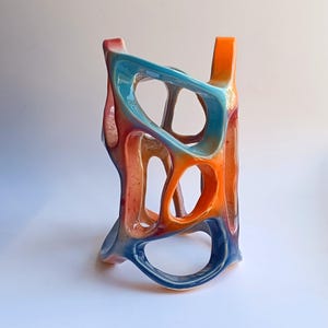 May include: A ceramic sculpture with a unique, abstract design. The sculpture is made of a light blue, orange, and red glaze. It has a hollow, open-work structure with multiple holes and openings.