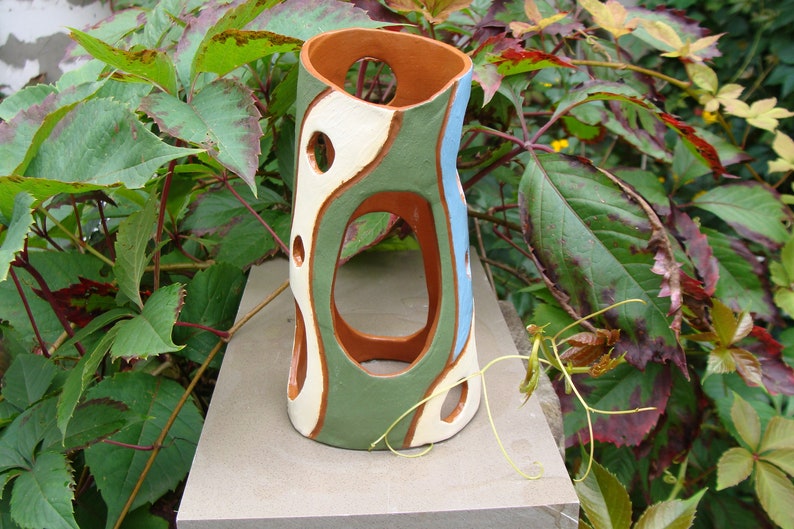 K&ouml;nnte beinhalten: Eine handgefertigte Keramikvase mit einem einzigartigen, offenen Design. Die Vase hat eine Mischung aus Farben, darunter Gr&uuml;n, Wei&szlig; und Blau, mit einem braunen Innenraum. Sie hat mehrere kreisf&ouml;rmige Aussparungen und ist etwa 25 cm hoch.