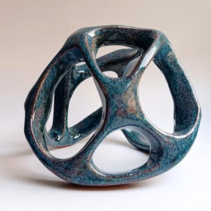 May include: A hollow, three-dimensional ceramic sculpture with a glossy, mottled finish in shades of deep teal, blue, and copper. The object features an open, interconnected structure with multiple rounded apertures and smooth, curved edges.