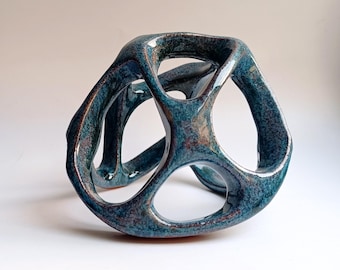 May include: A hollow, three-dimensional ceramic sculpture with a glossy, mottled finish in shades of deep teal, blue, and copper. The object features an open, interconnected structure with multiple rounded apertures and smooth, curved edges.