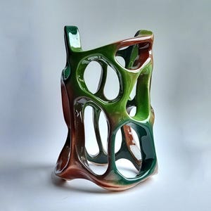 May include: A ceramic decorative vase with a unique, open design. The vase features a blend of colors, including shades of green and brown. The glossy finish and organic shape create a modern aesthetic. The vase has a height of approximately 10 inches.