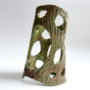 May include: A ceramic decorative vase with a tree bark texture. The vase is a light green color with brown accents and has several large, irregular holes. The vase is leaning slightly to the left.