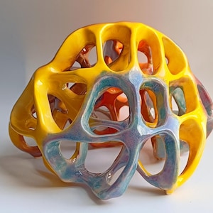 May include: A vibrant, abstract ceramic sculpture with a porous, organic design. The sculpture features a bright yellow exterior that transitions to shades of orange, blue, and purple within the openings. The artwork has a glossy finish and a unique, eye-catching form.
