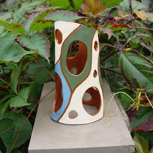 K&ouml;nnte beinhalten: Eine dekorative Keramikvase mit einzigartigem Design. Die Vase hat eine wei&szlig;e Basis mit braunen, gr&uuml;nen und blauen abstrakten Formen und kreisf&ouml;rmigen Ausschnitten. Die Vase ist etwa 25 cm hoch und steht auf einer Steinoberfl&auml;che, umgeben von gr&uuml;nem Laub.