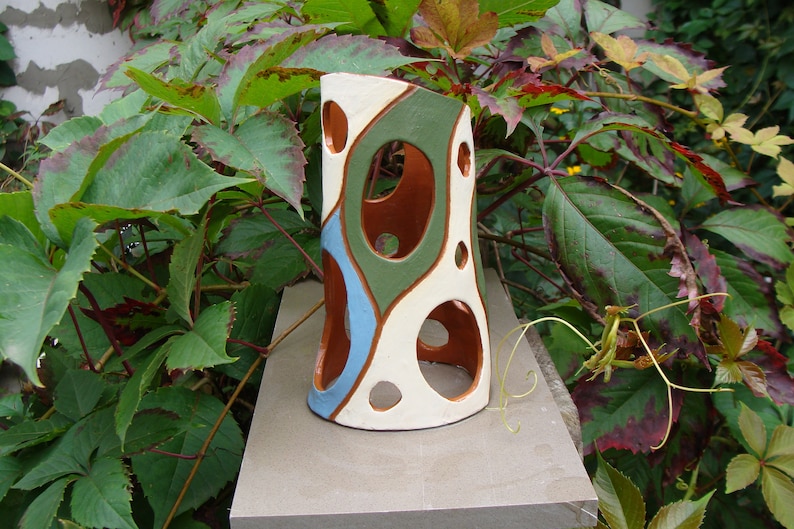 K&ouml;nnte beinhalten: Eine dekorative Keramikvase mit einzigartigem Design. Die Vase hat eine wei&szlig;e Basis mit braunen, gr&uuml;nen und blauen abstrakten Formen und kreisf&ouml;rmigen Ausschnitten. Die Vase ist etwa 25 cm hoch und steht auf einer Steinoberfl&auml;che, umgeben von gr&uuml;nem Laub.