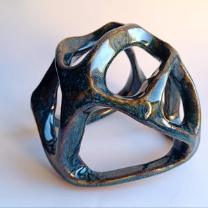 May include: A ceramic sculpture with a geometric, open design. The piece features a glossy, glazed finish with shades of blue, brown, and gold. The form is a continuous loop with multiple openings, creating a modern, abstract aesthetic.