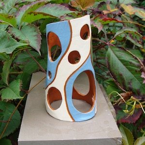 May include: A ceramic vase with a distinctive design. The vase has a light blue and white pattern with circular cutouts and brown accents. The cylindrical vase is on a stone surface, surrounded by green foliage.
