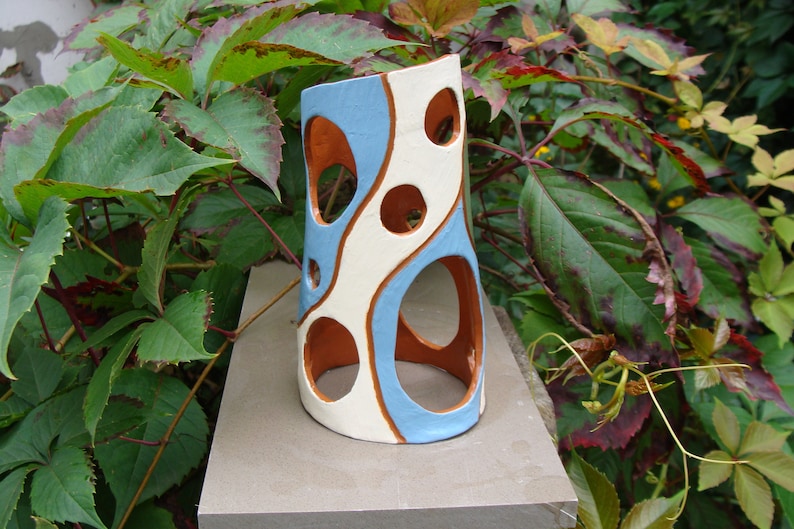 K&ouml;nnte beinhalten: Eine Keramikvase mit einzigartigem Design. Die Vase hat ein hellblaues und wei&szlig;es Muster mit kreisf&ouml;rmigen Aussparungen und braunen Akzenten. Die zylindrische Vase steht auf einer Steinoberfl&auml;che, umgeben von gr&uuml;nem Laub.