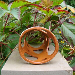 K&ouml;nnte beinhalten: Eine braune Keramikskulptur mit wei&szlig;em Innenraum und offenen, geometrischen Aussparungen. Die Skulptur befindet sich vor einem Hintergrund aus gr&uuml;nen und rot get&ouml;nten Bl&auml;ttern und ruht auf einer grauen Oberfl&auml;che. Das Kunstwerk hat eine kreisf&ouml;rmige Form.
