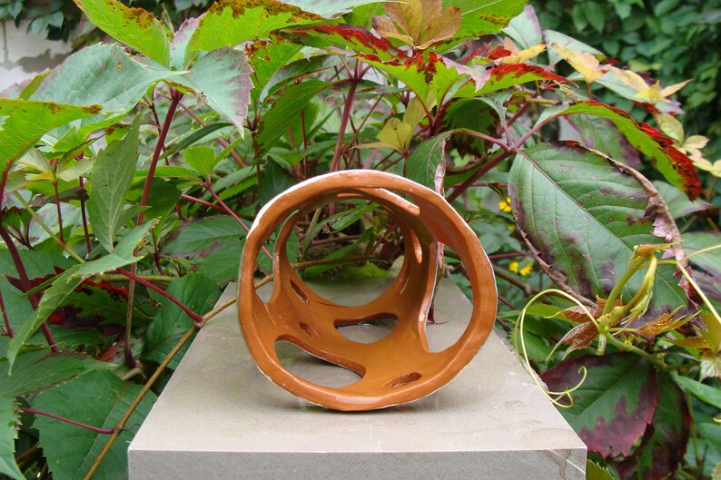 K&ouml;nnte beinhalten: Eine braune Keramikskulptur mit wei&szlig;em Innenraum und offenen, geometrischen Aussparungen. Die Skulptur befindet sich vor einem Hintergrund aus gr&uuml;nen und rot get&ouml;nten Bl&auml;ttern und ruht auf einer grauen Oberfl&auml;che. Das Kunstwerk hat eine kreisf&ouml;rmige Form.