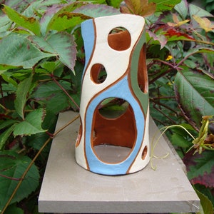 K&ouml;nnte beinhalten: Eine zylindrische Keramikvase mit einzigartigem Design. Die Vase hat eine cremefarbene Basis mit blauen, gr&uuml;nen und braunen organischen Formen und kreisf&ouml;rmigen Aussparungen. Die Vase wird im Freien vor einem Hintergrund aus gr&uuml;nem Laub gezeigt.