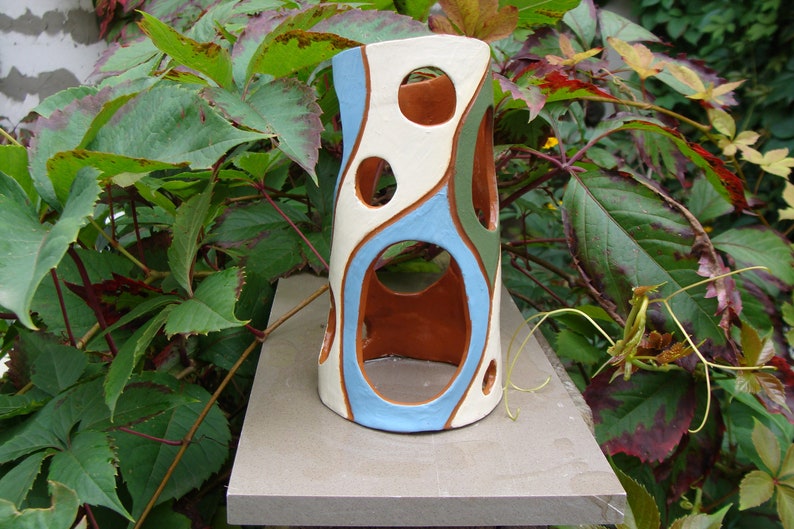 K&ouml;nnte beinhalten: Eine zylindrische Keramikvase mit einzigartigem Design. Die Vase hat eine cremefarbene Basis mit blauen, gr&uuml;nen und braunen organischen Formen und kreisf&ouml;rmigen Aussparungen. Die Vase wird im Freien vor einem Hintergrund aus gr&uuml;nem Laub gezeigt.