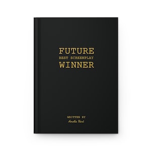 May include: A black hardcover journal featuring gold foil text on the front cover that reads FUTURE BEST SCREENPLAY WINNER, with the text WRITTEN BY Amelia Hart printed in smaller gold lettering near the bottom of the book cover.