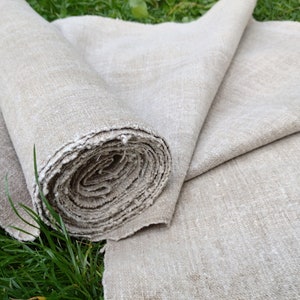 May include: A roll of natural-coloured linen fabric, partly unrolled, with a visible frayed edge. The fabric is lying on green grass, suggesting an outdoor setting. The linen has a textured appearance.