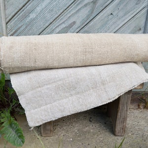 May include: Two rolls of natural linen fabric, one rolled and one flat, are placed on a small wooden stool. The fabric has a textured appearance and a light beige colour. The background features weathered, light blue wooden planks.