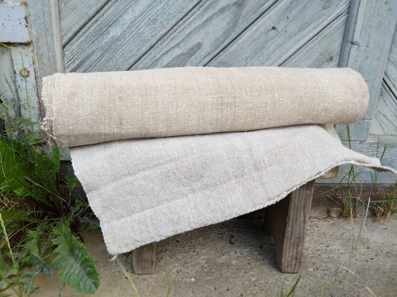 May include: Two rolls of natural linen fabric, one rolled and one flat, are placed on a small wooden stool. The fabric has a textured appearance and a light beige colour. The background features weathered, light blue wooden planks.