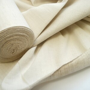 May include: A roll of natural-colored fabric, unbleached and woven, with a soft, textured surface. The fabric is folded and rolled, showing the raw edge.
