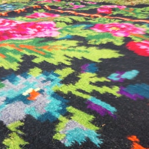 Op de afbeelding: Een kleurrijk geweven tapijt met een bloemenpatroon in tinten roze, groen, blauw en oranje op een zwarte achtergrond.