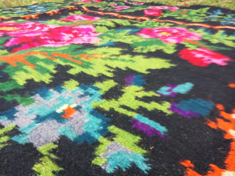 Op de afbeelding: Een kleurrijk geweven tapijt met een bloemenpatroon in tinten roze, groen, blauw en oranje op een zwarte achtergrond.