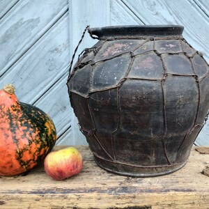 May include: A large, dark brown clay pot with a woven wire exterior. The pot is sitting on a wooden surface next to a small orange and green gourd and a red apple.