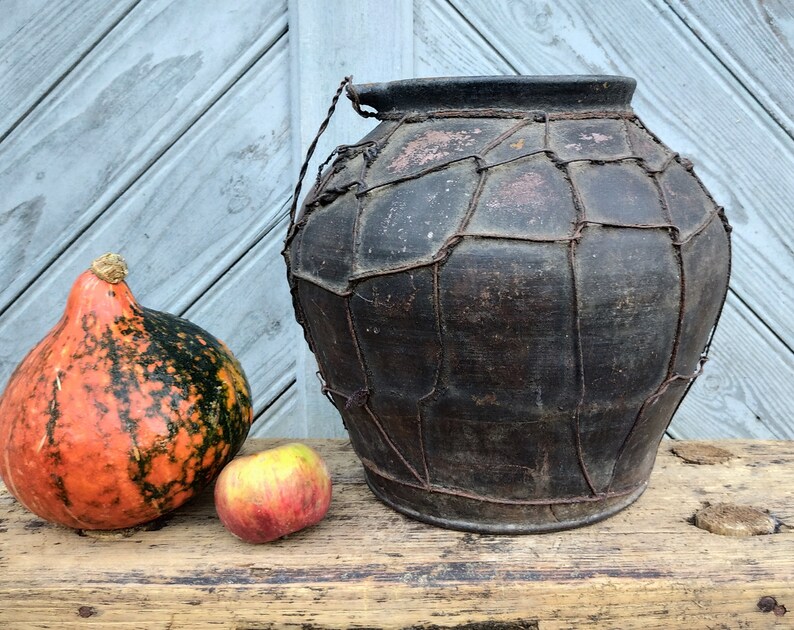 May include: A large, dark brown clay pot with a woven wire exterior. The pot is sitting on a wooden surface next to a small orange and green gourd and a red apple.