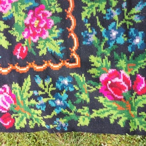Op de afbeelding: Close-up van een zwart wollen tapijt met een bloemenpatroon. Het tapijt heeft roze, rode, groene en blauwe bloemen op een zwarte achtergrond. Het tapijt heeft een gefranst randje.