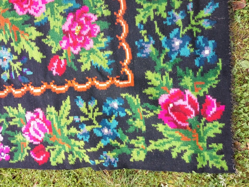 Op de afbeelding: Close-up van een zwart wollen tapijt met een bloemenpatroon. Het tapijt heeft roze, rode, groene en blauwe bloemen op een zwarte achtergrond. Het tapijt heeft een gefranst randje.