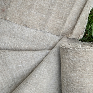 May include: Natural-coloured linen fabric, partly rolled, with a rough, textured weave. The fabric is a neutral beige colour, with visible fibres and a slightly frayed edge. The fabric is displayed on a green grassy surface.