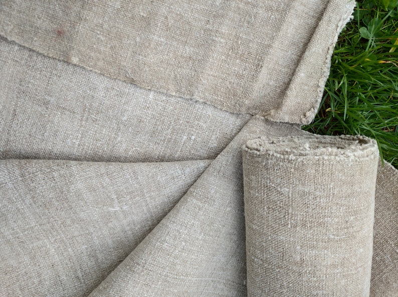 May include: Natural-coloured linen fabric, partly rolled, with a rough, textured weave. The fabric is a neutral beige colour, with visible fibres and a slightly frayed edge. The fabric is displayed on a green grassy surface.
