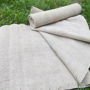 May include: Natural linen fabric, partly rolled and spread out on green grass. The fabric has a slightly textured appearance and a neutral, earthy tone. The rolled section suggests a length of fabric, while the unfolded part shows its width.
