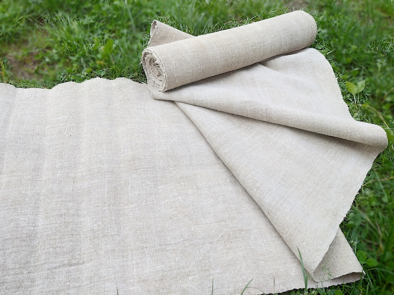 May include: Natural linen fabric, partly rolled and spread out on green grass. The fabric has a slightly textured appearance and a neutral, earthy tone. The rolled section suggests a length of fabric, while the unfolded part shows its width.