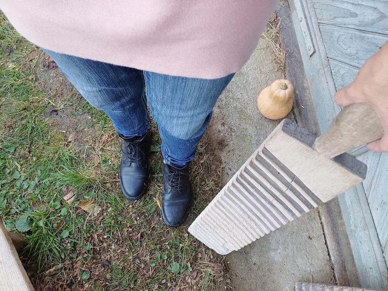 May include: A wooden washboard with a handle, sitting on a concrete surface. The washboard has a worn, rustic look and is made of light wood with dark grooves. A person is holding the handle of the washboard.
