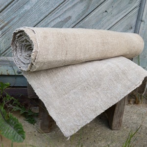May include: A rolled-up length of natural linen fabric sits on top of a folded piece of the same material. The fabric has a coarse, textured appearance, suggesting a rustic or vintage style. The neutral tones of the linen create a simple, earthy look.