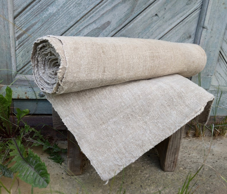 May include: A rolled-up length of natural linen fabric sits on top of a folded piece of the same material. The fabric has a coarse, textured appearance, suggesting a rustic or vintage style. The neutral tones of the linen create a simple, earthy look.